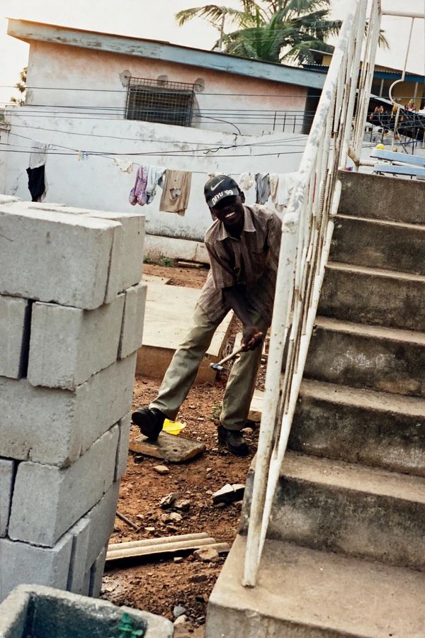 Kobe in Ghana in 1999, smiling with a hammer in hand beside a stack of concrete blocks and a staircase